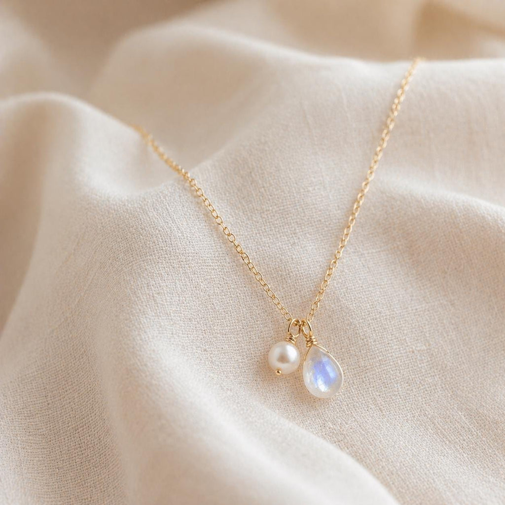 May include: A delicate gold necklace featuring a small, round pearl and a teardrop-shaped moonstone pendant. The necklace hangs on a fine gold chain, set against a soft, off-white fabric background. The jewelry is simple and elegant.