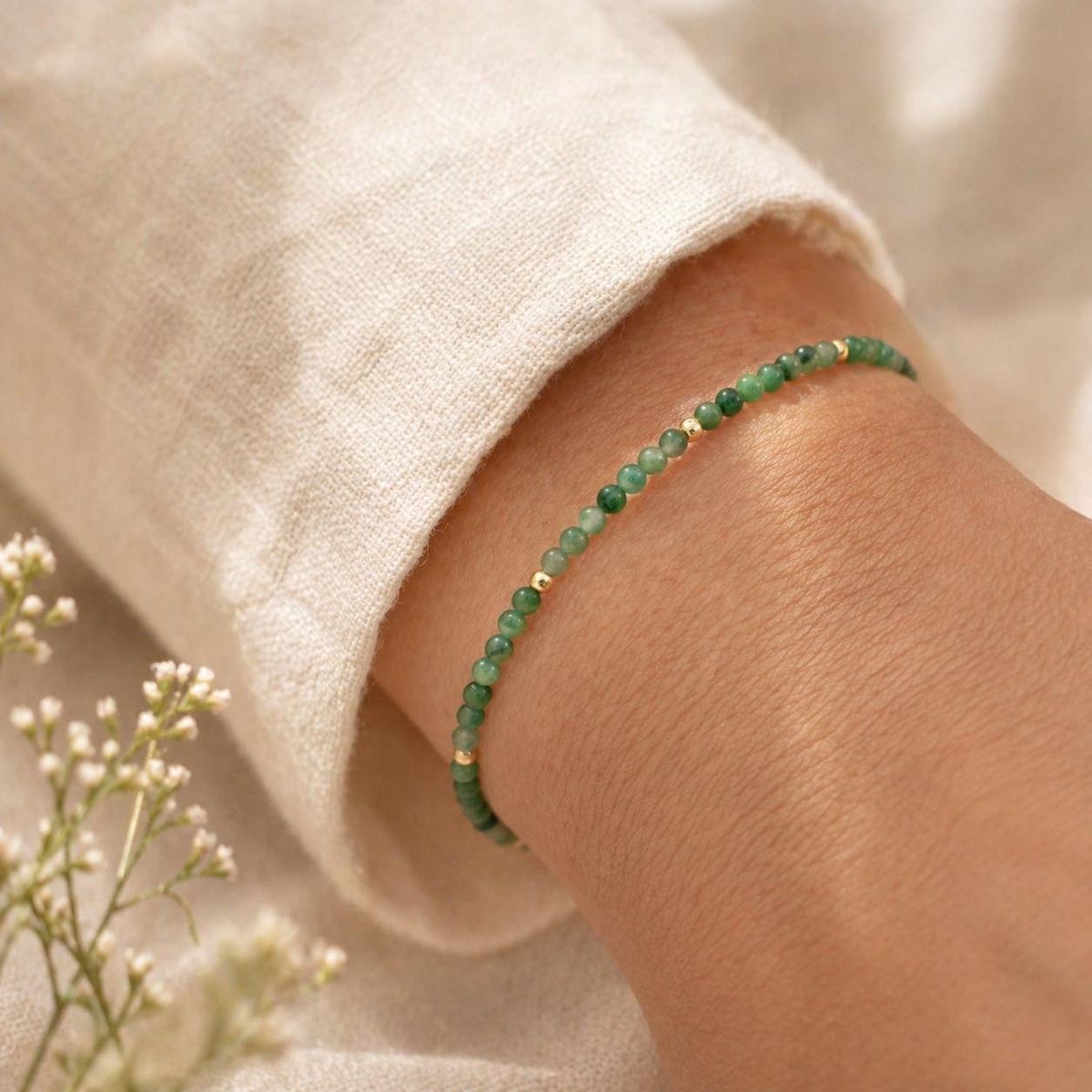 May include: A delicate bracelet featuring small, round green beads interspersed with gold-colored beads. The bracelet is worn on a wrist, with a cream-colored sleeve in the background. The bracelet is a simple, elegant piece of jewelry.